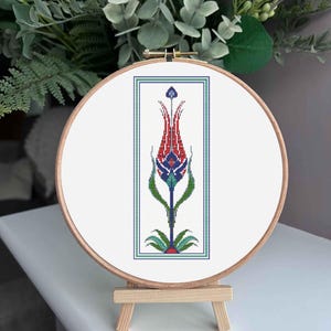 May include: A cross-stitch embroidery of a stylized red tulip with blue and green accents, framed in a rectangular border. The embroidery is displayed in a wooden hoop, set on a small wooden easel.