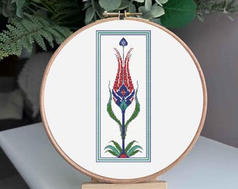 May include: A cross-stitch embroidery of a stylized red tulip with blue and green accents, framed in a rectangular border. The embroidery is displayed in a wooden hoop, set on a small wooden easel.