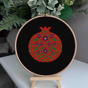 Peut inclure: Broderie au point de croix d'une grenade dans un cercle en bois. Le motif représente un fruit rouge avec des feuilles vertes et des fleurs bleues sur fond noir. La broderie est présentée sur un petit chevalet en bois.