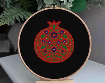 May include: A cross-stitch embroidery of a pomegranate in a wooden hoop. The design features a red fruit with green leaves and blue flowers on a black background. The embroidery is displayed on a small wooden easel.