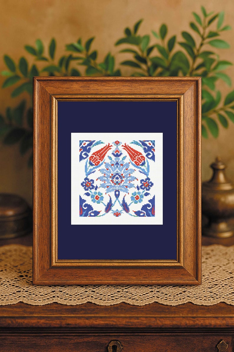May include: A framed cross-stitch artwork featuring a symmetrical floral design in red, blue, and white. The artwork is displayed in a wooden frame with a dark blue mat. The design includes stylized flowers and leaves.