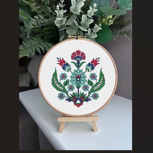 May include: A cross-stitch embroidery hoop featuring a floral design. The design incorporates red, blue and green flowers on a white background.