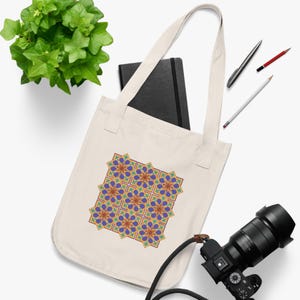May include: Cream-colored tote bag featuring a geometric floral pattern in shades of purple, blue, green, and gold. The bag has long straps and is made of a sturdy fabric. A black notebook, pens, and a camera are also visible.
