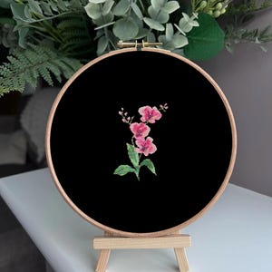 May include: A pink flower embroidered on black fabric in a wooden embroidery hoop on a wooden easel.