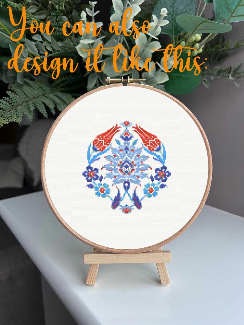 May include: A cross-stitch embroidery hoop featuring a floral design in red, blue, and white. The design is centered on a white fabric, held within a wooden hoop. The text "You can also design it like this" is in orange script.