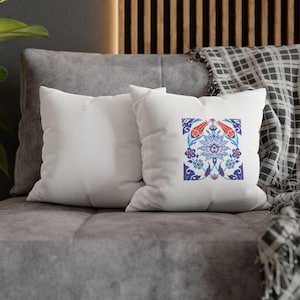 May include: Two white square throw pillows with a colorful floral design. The design features red, blue, and white flowers and leaves. The pillows are on a gray couch with a patterned blanket.