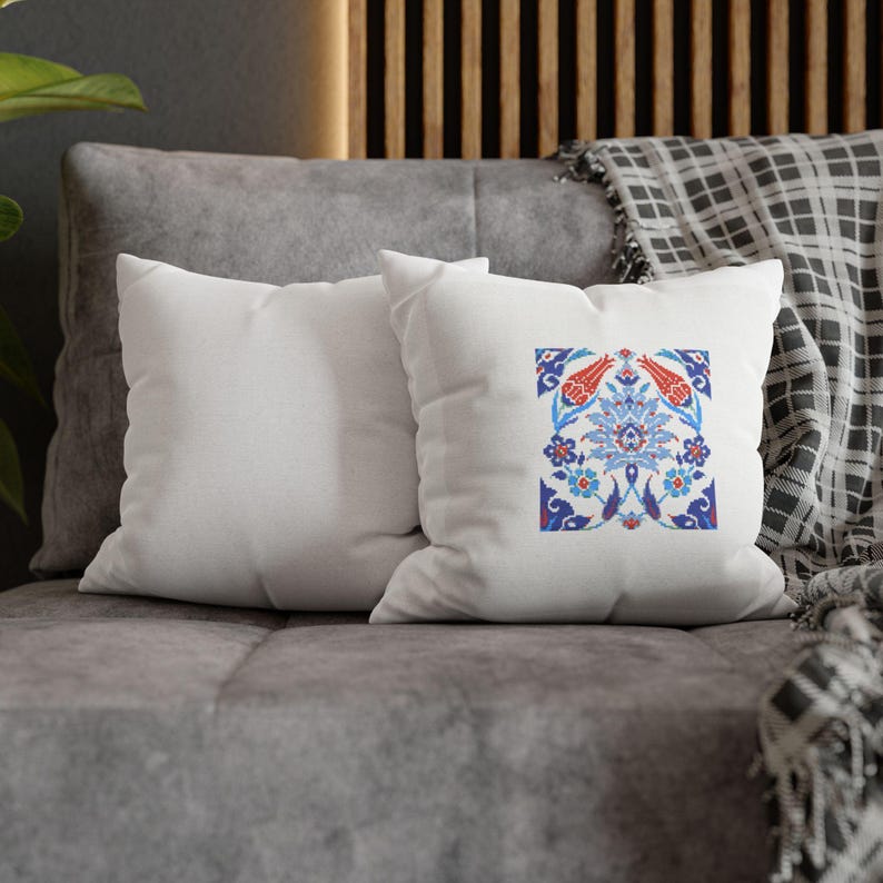 May include: Two white square throw pillows with a colorful floral design. The design features red, blue, and white flowers and leaves. The pillows are on a gray couch with a patterned blanket.