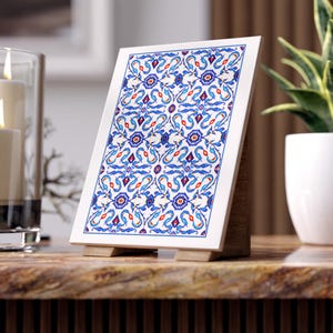 May include: A decorative white tile with a vibrant blue, red, and white floral pattern, displayed on a wooden easel. The tile features an intricate design with symmetrical elements, creating a visually appealing piece for home decor.