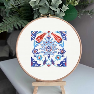 May include: A cross-stitch embroidery hoop featuring a detailed floral design in red, blue, and white. The design includes stylized flowers and geometric patterns, set against a white background. The hoop is displayed on a small wooden easel.