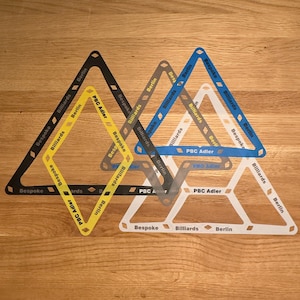 May include: A selection of coloured billiard triangle racks. The racks are in black, yellow, grey, blue, and white. Each rack has the words "Bespoke", "Billiards", "Berlin", and "PBC Adler" printed on them. The racks are arranged on a wooden surface.