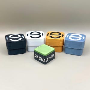 Puede incluir: Una colección de tizas cuadradas en varios colores: negro, blanco, naranja y azul. Cada soporte tiene un logotipo "e" blanco. Un cubo de tiza verde y negro con el texto "PAGULAYAN" está al frente.