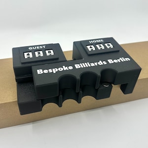 Peut inclure: Dispositif de pointage noir avec un compteur numérique pour les joueurs à domicile et les invités. L'appareil est étiqueté "Bespoke Billiards Berlin".