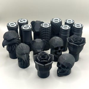 May include: A collection of black decorative bottle stoppers. The stoppers feature various designs, including an eagle head, bear head, skull, rose, and cylindrical shapes with a white logo. The stoppers are made of a matte black material.