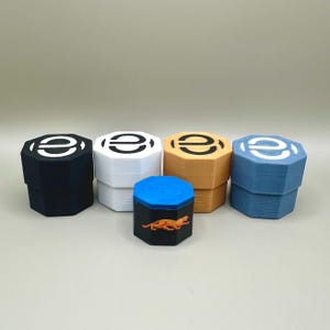 Könnte beinhalten: Fünf kleine, achteckige Behälter mit Deckeln. Vier Behälter haben ein weißes Logo mit einem schwarzen Kreis darin. Ein Behälter hat einen schwarzen Deckel mit einer blauen Oberseite und einem kleinen orangefarbenen Eidechsen-Design.