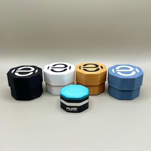 以下が含まれることがあります： 黒と白のロゴが上部に付いた、小さな六角形の容器が5つあります。容器は黒、白、茶色、青、ターコイズです。ターコイズの容器には、側面に「PURE」と印刷されています。