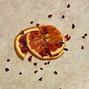 May include: Two dried orange slices with a reddish-brown hue, adorned with small, dark red and brown pieces. The orange slices are arranged on a light-colored surface, creating a natural and rustic aesthetic.