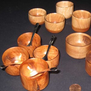 May include: A set of eight small wooden bowls with a natural wood grain finish. The bowls are arranged in a semi-circle on a dark surface. Each bowl has a small silver spoon inside.