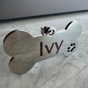 May include: A silver-coloured dog tag shaped like a bone. The tag features the name "Ivy" in a dark red font and a paw print. It has a small loop for attaching to a collar.