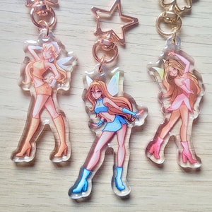 Puede incluir: Tres llaveros acrílicos con personajes de hadas de dibujos animados en varias poses. Cada llavero tiene un cierre dorado y un dije con un hada con un atuendo diferente. Las hadas visten atuendos en tonos naranja, azul y rosa.