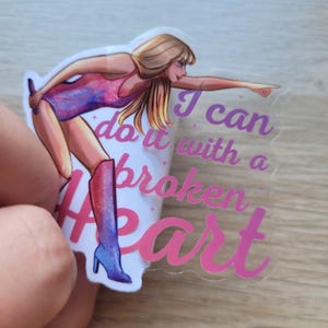 May include: A clear sticker with a pink and purple design. The sticker features a cartoon woman with long blonde hair wearing a pink and purple outfit and boots. The text on the sticker reads "I can do it with a broken heart".