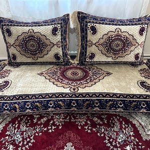 Puede incluir: Un cojín de suelo de color crema con un borde estampado en azul y rojo. Dos almohadas con un patrón similar se colocan encima del cojín.