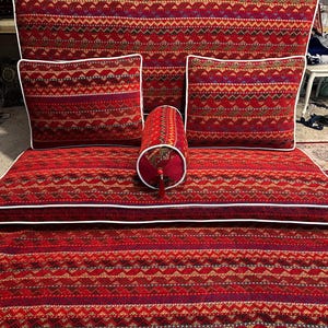 Puede incluir: Un cojín de suelo rojo y blanco con un borde blanco. El cojín está hecho de tela y tiene un diseño geométrico. Hay tres almohadas en el cojín, dos rectangulares y una cilíndrica. Las almohadas también son rojas y blancas con patrones.