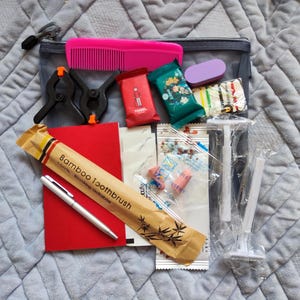 Puede incluir: Una colección de artículos de aseo y accesorios de viaje, que incluyen un peine rosa, abrazaderas negras, un bloc de notas rojo, un bolígrafo, un cepillo de dientes de bambú y jabones envueltos. Los artículos están dispuestos sobre una superficie gris.