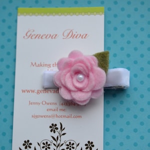 Peut inclure: Une pince à cheveux en forme de fleur en feutre rose avec une base blanche et des feuilles en feutre vert. La pince est sur une carte qui dit "Geneva Diva" et "Making the ordinary extraordinary".
