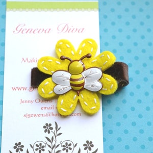 May include: A yellow felt flower with a brown felt backing and a bee in the center. The bee is made of plastic and has black and yellow stripes. The flower has white stitching around the edges.