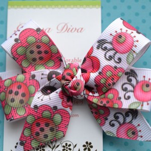 May include: A white grosgrain ribbon hair bow featuring a repeating pattern of stylised pink and green flowers with dark brown centres. The bow is tied in a central knot and is displayed on a white rectangular card with partial text visible.