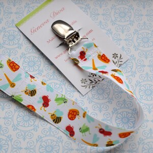 May include: White pacifier clip with a silver clip and a white ribbon with a pattern of colorful bugs, including ladybugs, bees, and green insects. The ribbon is attached to the clip. The clip is labeled "Geneva Diva".