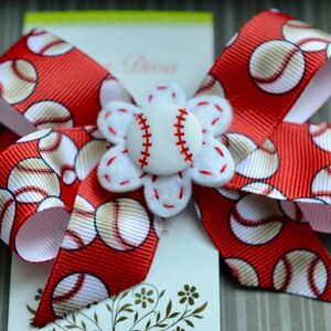 May include: A red and white baseball-themed hair bow with a white felt flower and a white baseball button in the center.
