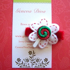 May include: A white felt flower hair clip with a red and green glitter center, red stitching, and a red velvet band. The business card behind the clip reads "Geneva Diva" and "Bows are your passion".