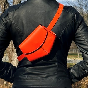 May include: A bright orange leather fanny pack with a curved shape and a black adjustable strap. The fanny pack is worn by a person wearing a black leather jacket.