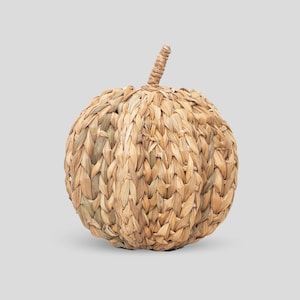 May include: A decorative pumpkin-shaped object crafted from woven light brown natural seagrass fibres. The item features a rounded, segmented body with a prominent, upright stem at the top, showcasing a detailed braided texture throughout the surface.