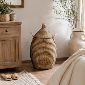 May include: A tall, oval-shaped woven basket with a dark brown finish, featuring a matching lid with a small knob handle on top. The basket includes two thick, light-coloured rope handles attached to the sides for lifting and carrying.
