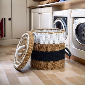 Op de afbeelding: Een ronde gevlochten wasmand met een afneembaar deksel, voorzien van horizontale banden van natuurlijk bruin, wit en zwart zeegras. De mand heeft een wit touw handvat op het deksel en een zwart touw handvat aan de zijkant voor transport.