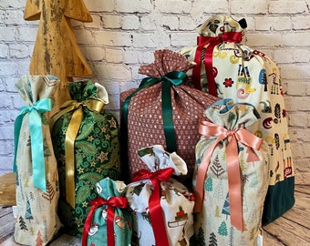 May include: A collection of seven Christmas gift bags made of fabric with various holiday patterns. The bags are decorated with ribbons in red, green, and gold. The bags are arranged on a wooden surface in front of a white brick wall.