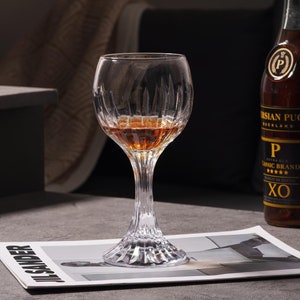 Könnte beinhalten: Ein klares Glas mit geriffeltem Design, gefüllt mit bernsteinfarbener Flüssigkeit, steht auf einem Magazin. Das Glas hat einen langen Stiel und eine runde Schale. Eine Flasche XO Brandy ist im Hintergrund zu sehen.