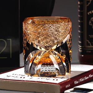 May include: An amber-coloured cut crystal whiskey glass with a geometric pattern. The glass sits on a book titled "What I Wish I Knew When I Wish". The glass has a thick base and a detailed, textured design. The book is in English.