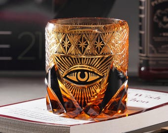 May include: A gold and black glass tumbler with an all-seeing eye design. The glass has a geometric pattern and is perfect for serving cocktails or whiskey.