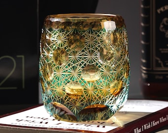 Könnte beinhalten: Ein dekorativer, geschliffener Glasbecher mit geometrischem Muster. Das Glas hat einen Farbverlauf, der von Goldgelb oben nach Türkisgrün unten übergeht. Der Becher steht auf einem Buch.