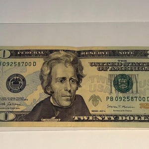 May include: A twenty-dollar bill featuring a portrait of Andrew Jackson. The bill is primarily green and black, with the text "Federal Reserve Note" and "Twenty Dollars" prominently displayed. The serial number is PB 09258700 D.