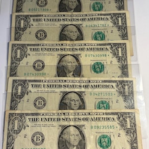 May include: Five United States one-dollar bills are displayed, each featuring a portrait of George Washington. The bills are in clear protective sleeves. The text "THE UNITED STATES OF AMERICA" is visible on each bill, along with the denomination "ONE DOLLAR".