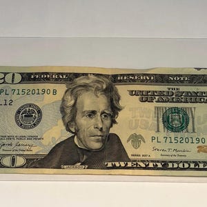 May include: A twenty-dollar bill in a clear plastic sleeve. The bill features a portrait of Andrew Jackson and the text "TWENTY DOLLARS". The bill is in good condition.