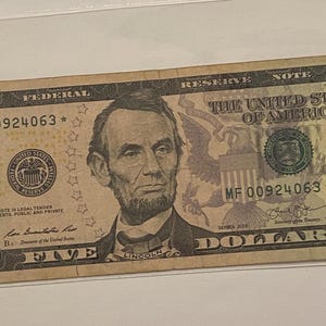 May include: A United States five-dollar bill. The bill features a portrait of Abraham Lincoln and the text "The United States of America". The bill is tan and green in color.