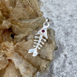 May include: Silver fish bone charm with a red gemstone eye. The charm is hanging from a silver loop.