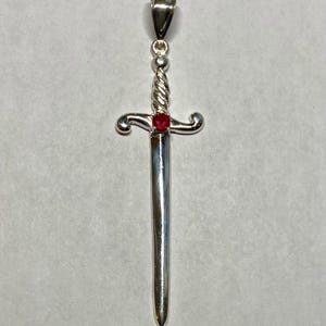 Puede incluir: Un colgante de espada de plata con una piedra preciosa roja engastada en la empuñadura.