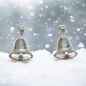 Op de afbeelding: Twee zilveren belhangers met edelsteenaccenten. Eén bel heeft een groene edelsteen, de andere een roze edelsteen. De bellen staan tegen een besneeuwde achtergrond, wat een feestelijk thema suggereert.