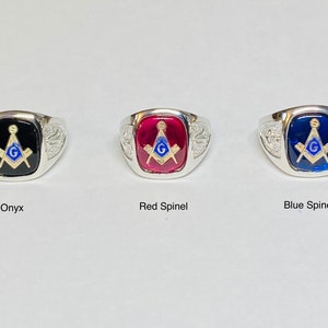 May include: Three silver Masonic rings with different colored stones. The first ring has a black onyx stone, the second ring has a red stone, and the third ring has a blue stone. Each ring features a gold Masonic symbol with a blue square and compass.